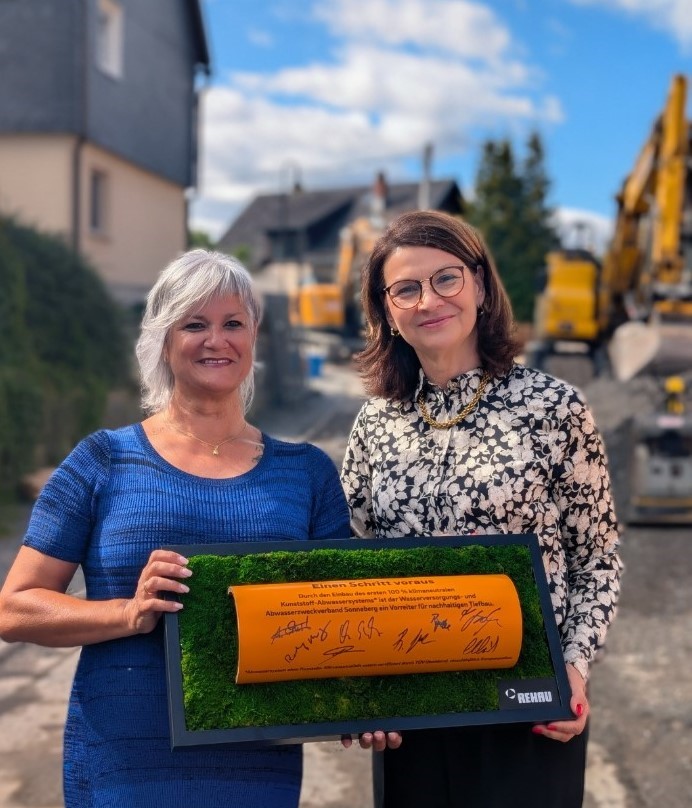 Im Bild sieht man links die Leiterin Abwasser der Wasserwerke, Frau Ernst, und rechts im Bild Frau Große, Vertreterin der Firma Rehau. Sie beide halten das eingerahmte und unterschriebene Stück Kanalrohr in der Hand. Sie lächeln freundlich i die Kamera.
