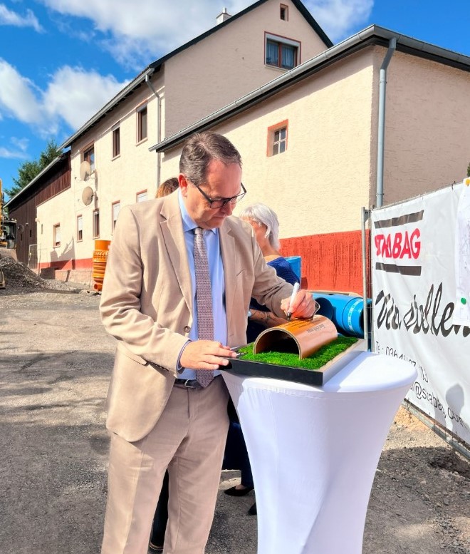 IM Bild sieht man den Werkleiter der Wasserwerke, Herrn Hubner. Er unterschreibt auf einem Stück des klimaneutralen Kanalrohrs, welches als Bild eingerahmt wurde.