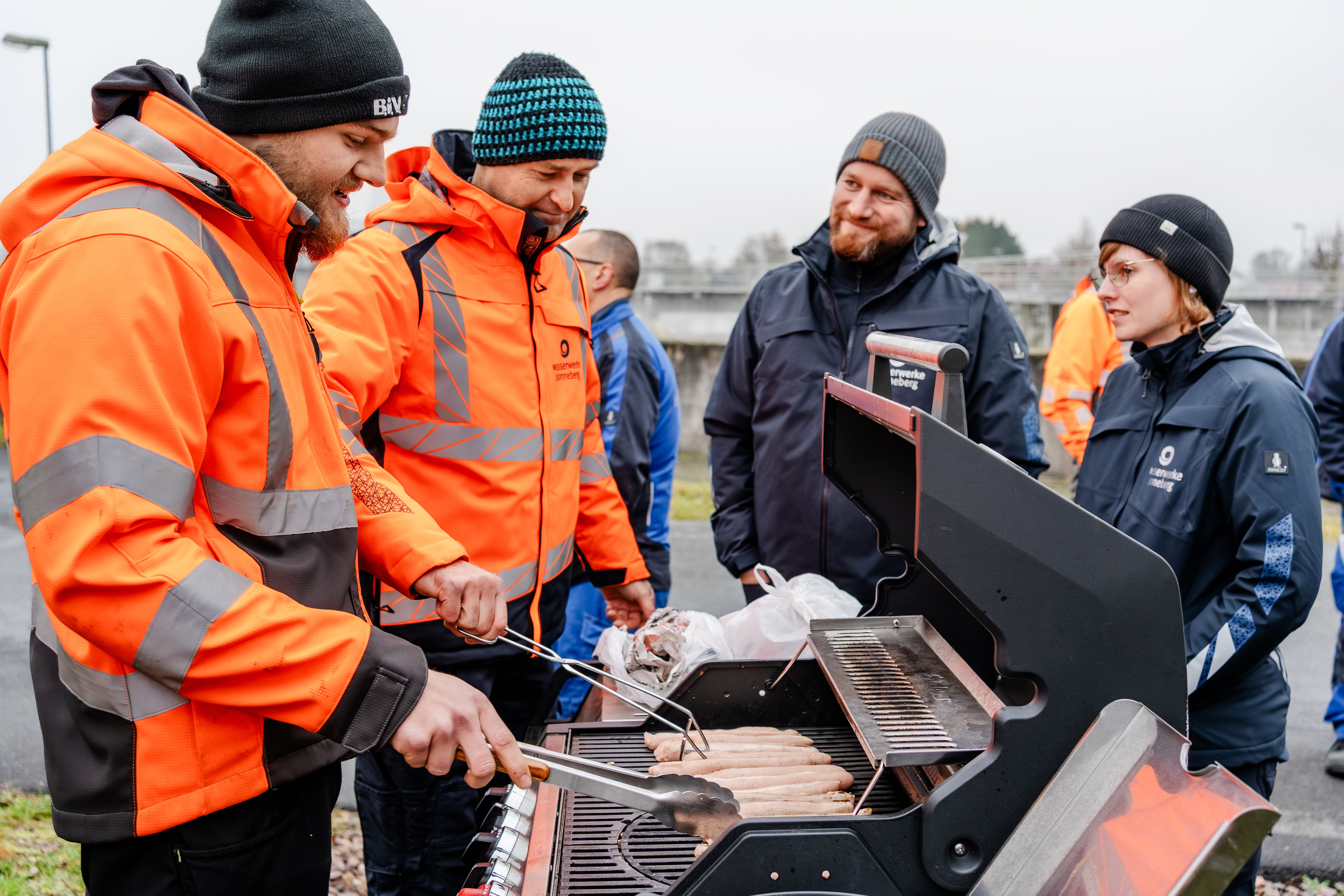 Im Bild sieht man drei Mitarbeiter und eine Mitarbeiterin der Wasserwerke Sonneberg. Sie stehen gut gelaunt um einen Grill herum. Zwei der Mitarbeiter kümmern sich um die Brautwürste darauf. Sie alle haben dicke Arbeitsjacken an und eine Mütze auf.